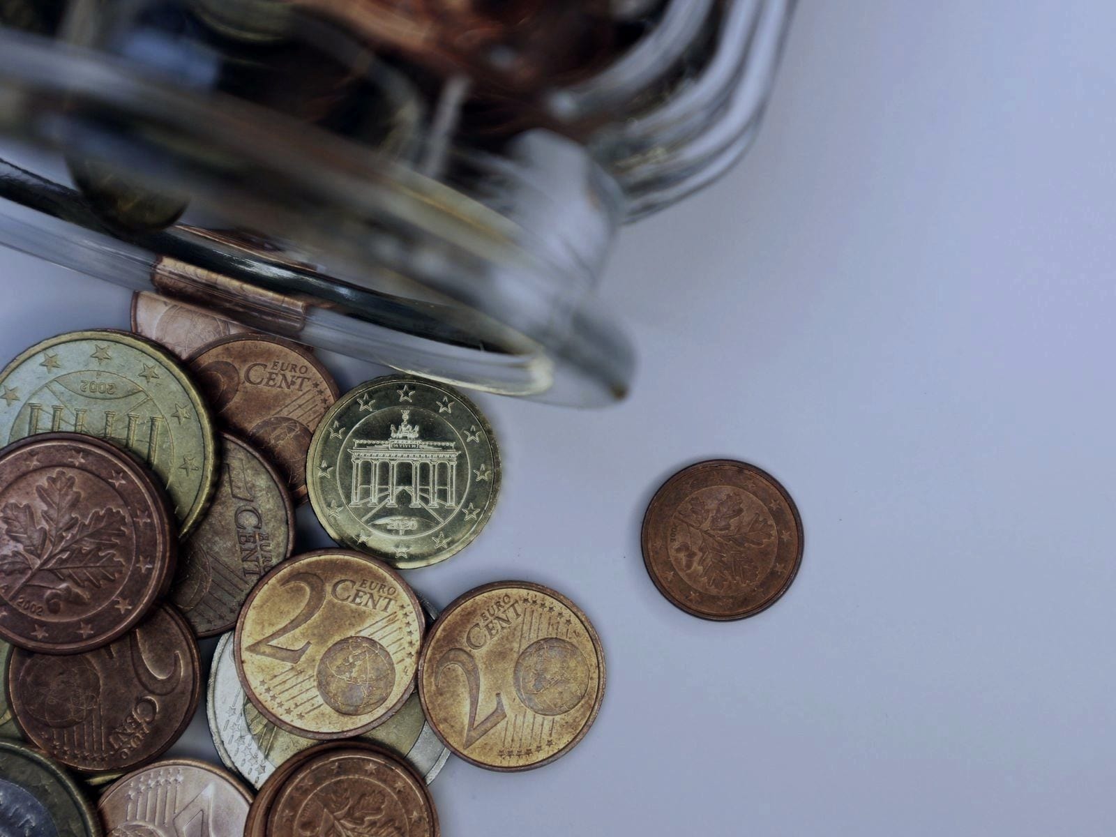 Moedas de euro de vários valores espalhadas sobre uma superfície branca, a sair de um frasco de vidro.