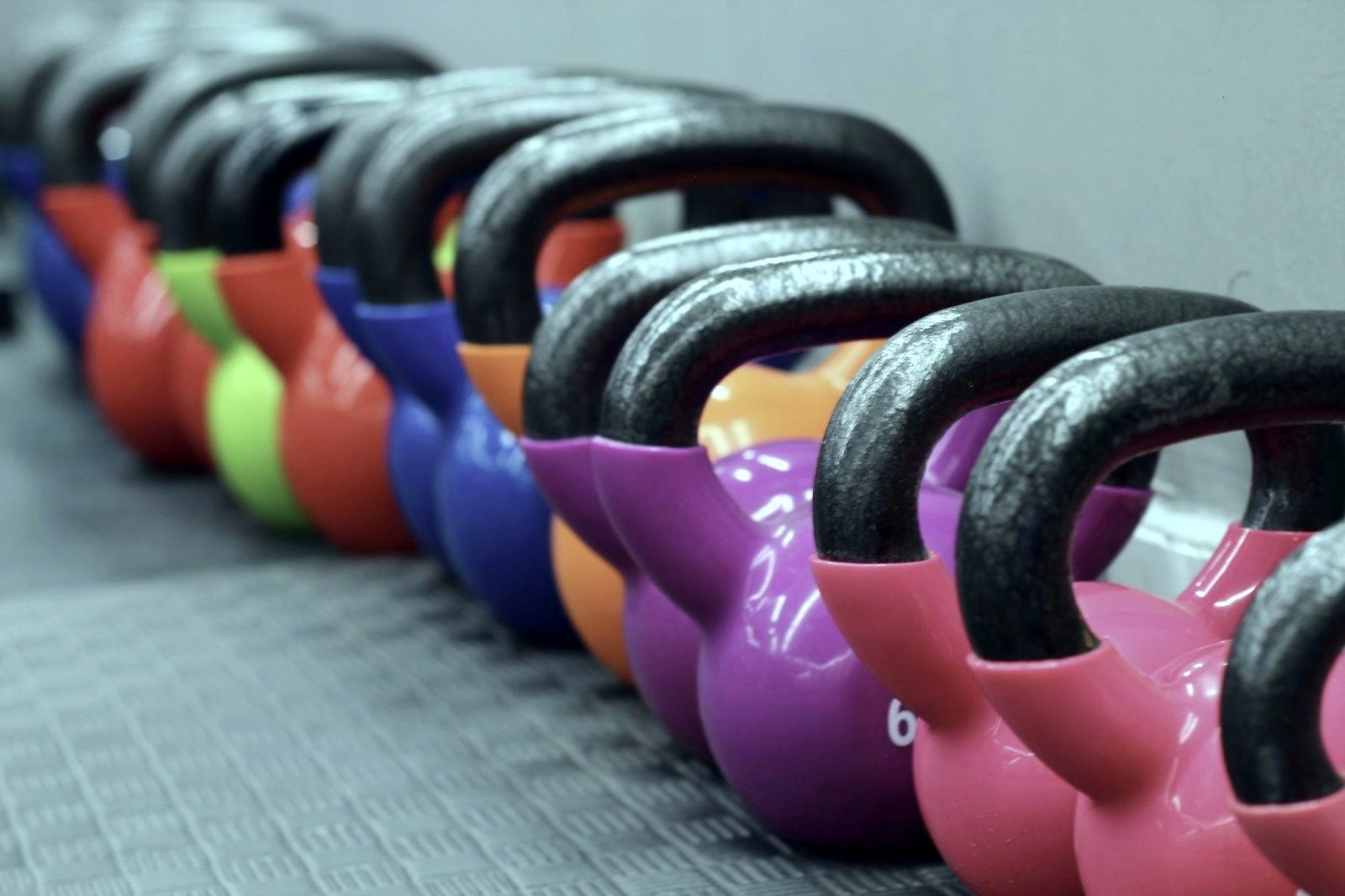 Fila de kettlebells de várias cores e pesos alinhadas no chão de um ginásio.